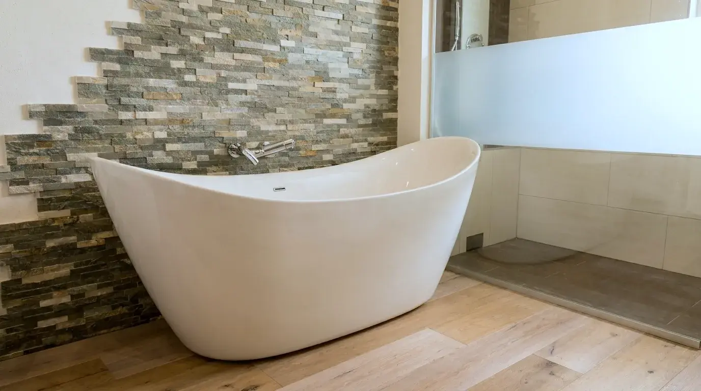 Freistehende Badewanne mit hoher Rückenlehne vor einer Wand aus gestapelten Natursteinriemchen, rechts eine bodengleiche Dusche mit beigen Großformatfliesen, Boden aus breiten Eichendielen