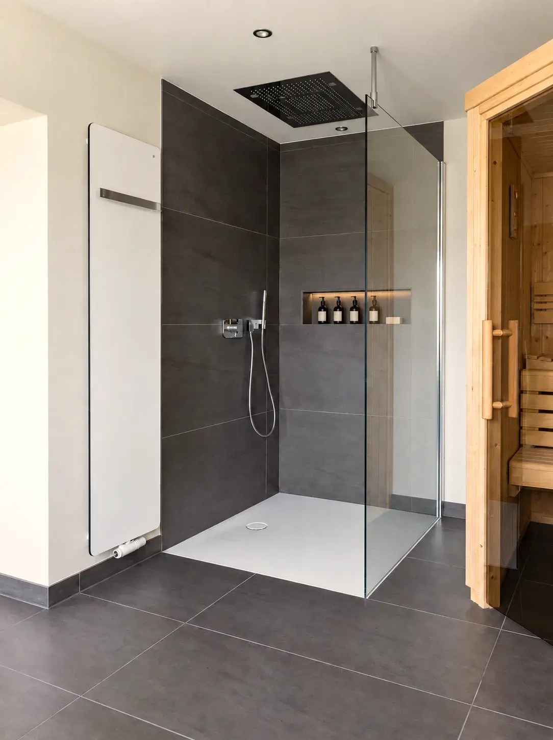 Walk-in-Dusche mit großer Deckenregenbrause und fester Glasabtrennung, links ein Handtuchheizkörper, rechts die Sauna mit Glastür