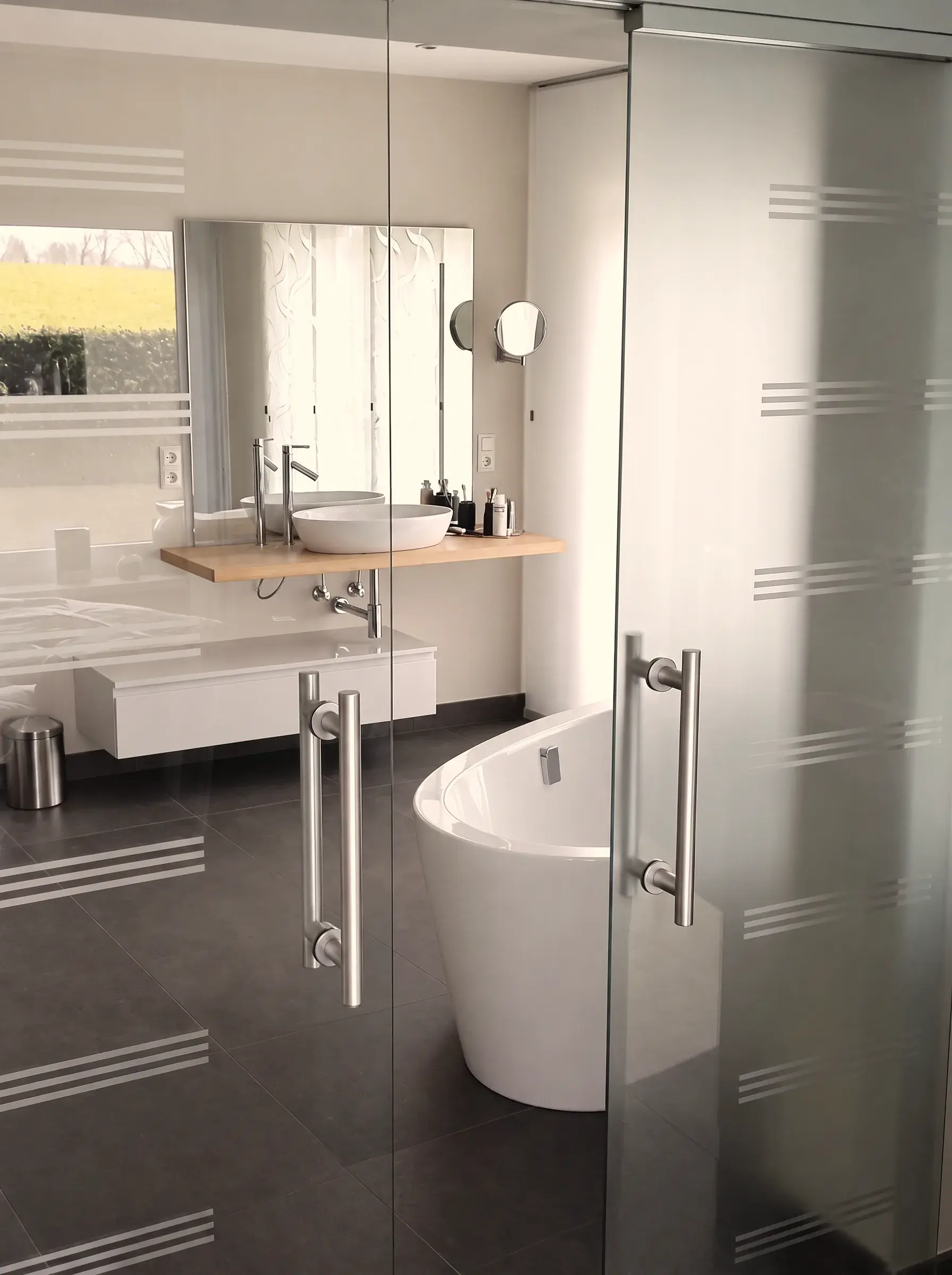 Blick durch die zweiflügelige Glasschiebetür ins Bad: Waschtisch mit Aufsatzbecken auf einer Eichenablage, großer rahmenloser Spiegel und die freistehende Wanne